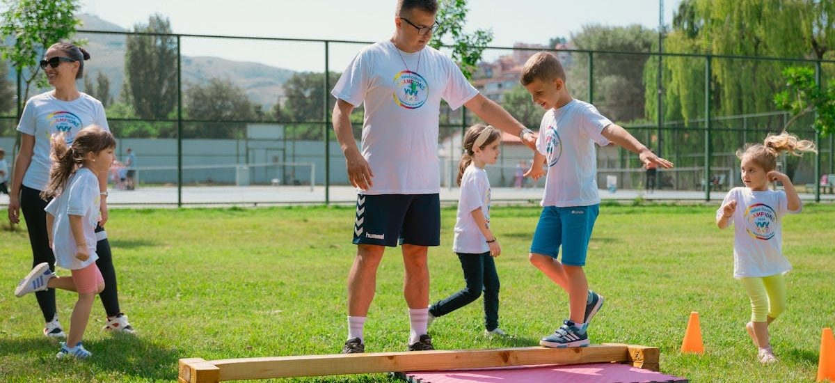 Kids guided by instructors at a park balance on a beam for fun and exercise.