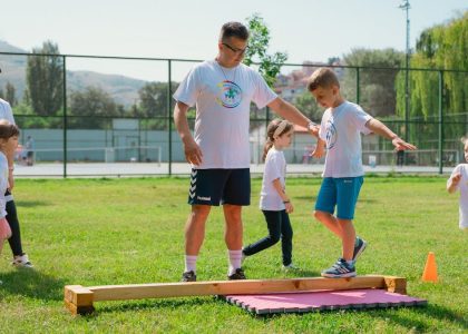 Kids guided by instructors at a park balance on a beam for fun and exercise.
